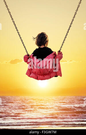 Bambina sull'altalena guardando il tramonto sul mare Foto Stock