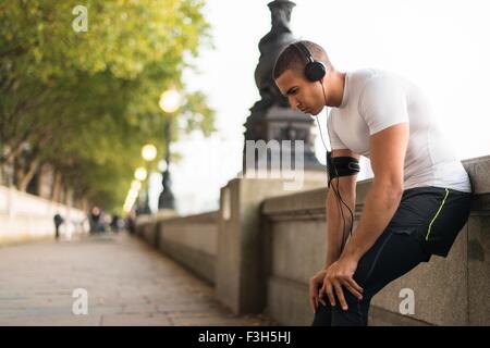 Giovane maschio runner indossa le cuffie prendendo una pausa su Riverside Foto Stock