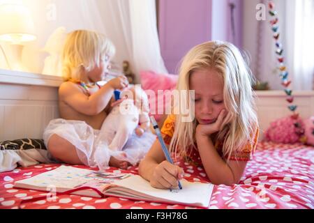 Ragazza e la sorella toddler giacente sul letto giocando con il libro da colorare Foto Stock