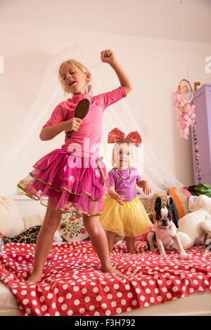 Ragazza e la sorella toddler danzando e cantando con microfono sul letto Foto Stock