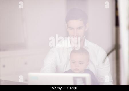 Padre utilizzando laptop mentre il giovane figlio siede sul suo giro, presa attraverso la finestra Foto Stock