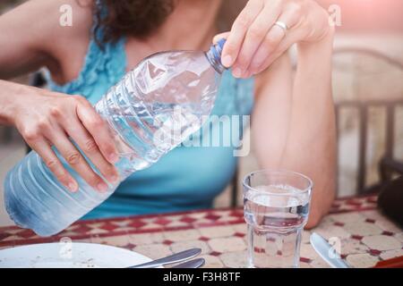 Ritagliato colpo di donna matura versando un bicchiere di acqua in bottiglia a tavola Foto Stock