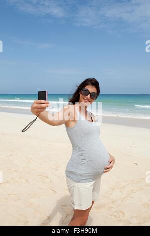 Gravidanza metà donna adulta prendendo selfie smartphone sulla spiaggia, Capo Verde, Africa Foto Stock