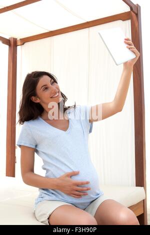 Gravidanza metà donna adulta seduto sul divano letto prendendo tavoletta digitale selfie Foto Stock