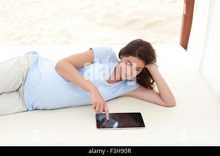 Gravidanza metà donna adulta che giace sulla spiaggia di dormeuse con tavoletta digitale Foto Stock