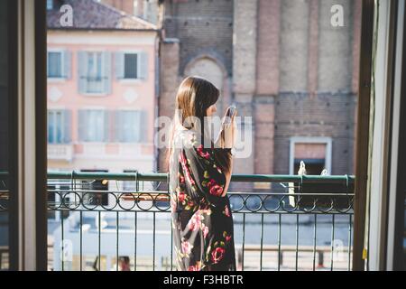 Giovane donna la lettura di testi di smartphone sul balcone della città Foto Stock