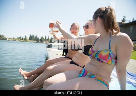 Giovani donne che indossano bikini tostare con la birra sul molo fronte mare, Lake Oswego, Oregon, Stati Uniti d'America Foto Stock