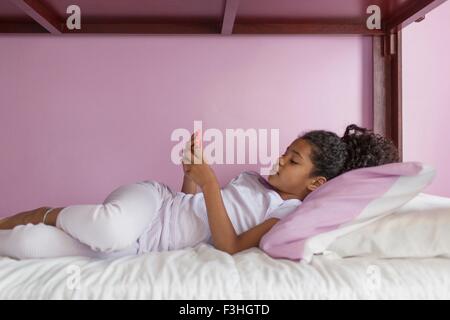 Vista laterale della ragazza indossando pigiami giacente sul letto guardando smartphone Foto Stock