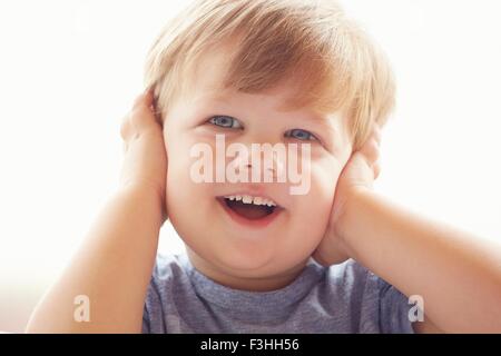 Ritratto di giovane ragazzo che copre le orecchie con le mani, guardando lontano sorridente Foto Stock