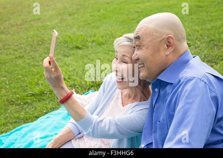 Coppia senior seduta nel parco, tenendo autoritratto utilizza lo smartphone Foto Stock