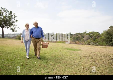 Coppia senior camminare a braccetto nella park, portando un cestino picnic Foto Stock