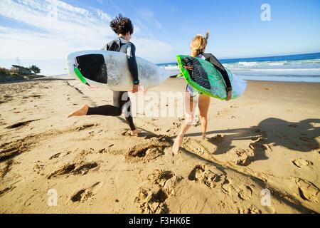 Giovane che corre verso il mare, portando le tavole da surf, vista posteriore Foto Stock