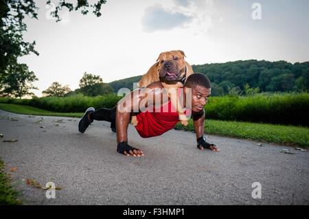 Giovane uomo che fa pushups sulla strada rurale offrendo nel contempo cane un piggyback Foto Stock