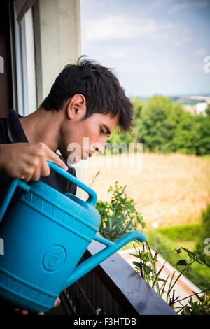 Attraente giovane uomo su Appartamento balcone Impianti di irrigazione nella casella dal blu Annaffiatoio sulla giornata di sole con il campo in background Foto Stock