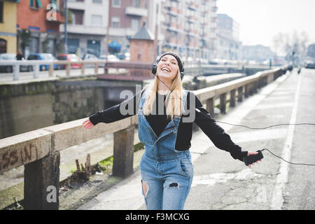 Ginocchio la figura del giovane bella bionda capelli dritti donna della città con le cuffie per ascoltare musica, braccia spalancate, sentendosi liberi, ridendo - la libertà, il concetto di felicità Foto Stock