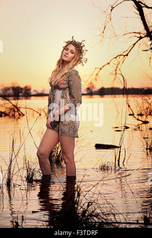 Una giovane donna che indossa una copertura, costume da bagno e un fiorito vetch di godersi la natura mentre guadare in un lago al tramonto. Foto Stock