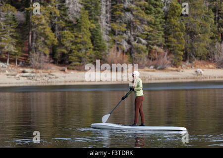 Un attivo, giovane donna vestita di una felpa e un cappello di maglia paddling una racchetta di bordo su un piccolo lago di montagna in California. Foto Stock