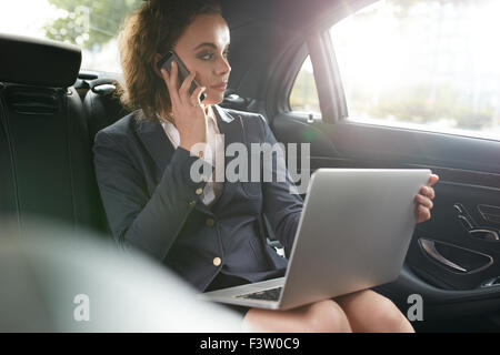 Imprenditrice seduta sul sedile posteriore della vettura con il computer portatile parlando al telefono cellulare. Executive femmina che viaggiano per lavoro in auto di lusso Foto Stock