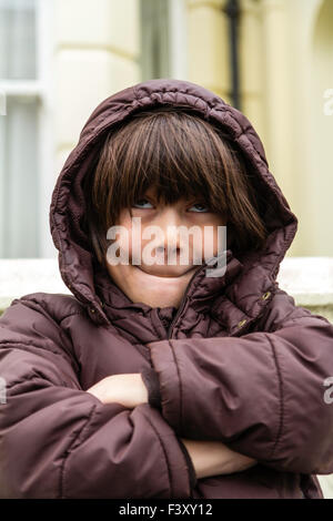 Dieci anni ragazzo caucasico in piedi fuori, freddo, braccia incrociate, in giacca a vento, tirando comico e faccia sorridere mentre guardando verso l'alto Foto Stock