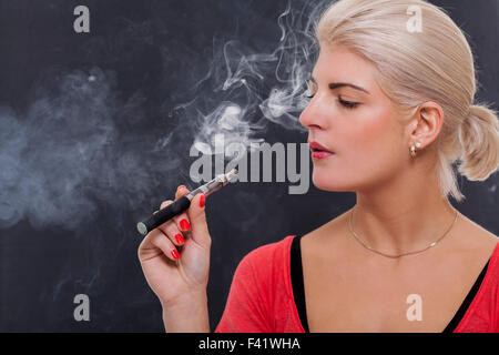 Elegante donna bionda di fumare un e-sigaretta espirando una nube di fumo con gli occhi chiusi in godimento, vista di profilo su uno sfondo scuro Foto Stock