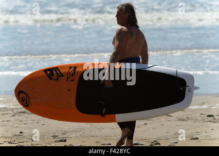 Il surfer che trasportano stand up paddle board giù in spiaggia per la scintillante white surf su Gower, Wales, Regno Unito Foto Stock