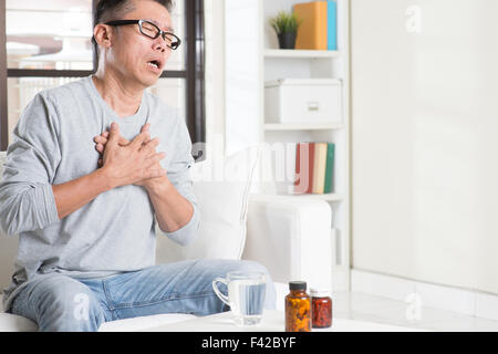 Coppia 50s uomo asiatico heartache, premendo sul petto con espressione dolorosa, seduto sul divano di casa, le medicine su tavola. Foto Stock