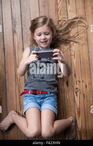 Bambina giacente sul piano utilizza lo smartphone Foto Stock