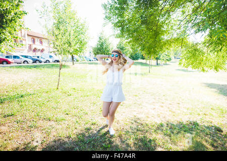 Giovani caucasici belli lunghi biondi capelli dritti hippy woman dancing in un parco cittadino, divertendosi, mani dietro la testa, toccando i capelli - musica, danza, concetto di felicità Foto Stock