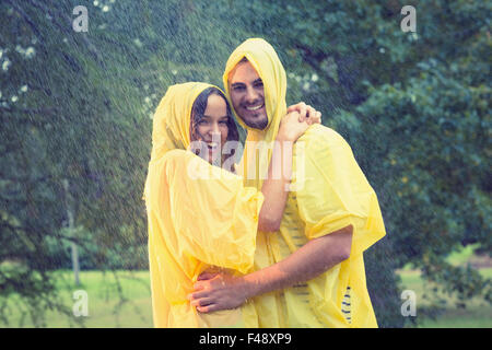 Carino coppia indossando protezione capo e abbracciando sotto la pioggia Foto Stock