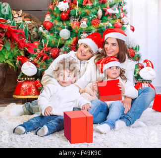 Allegro madre con i suoi tre bambini preziosi rosso da indossare cappelli di Babbo Natale seduto vicino al bellissimo albero di Natale decorato Foto Stock
