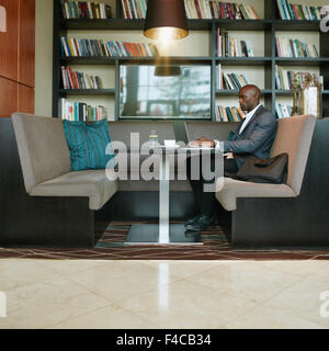 Colpo di giovane imprenditore africano seduto da solo presso la caffetteria al lavoro sul suo computer portatile. In attesa del cliente per una riunione. Foto Stock