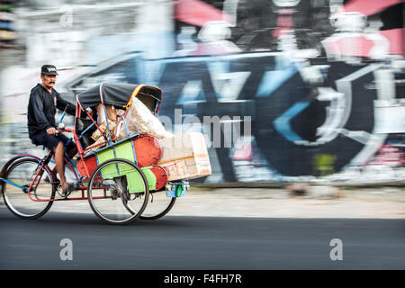 Risciò ciclo con passeggero accelerando lungo in questa fotografia panoramica Scena di strada di città indonesiane la vita con colorati graffiti sullo sfondo della parete Foto Stock