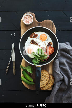 Pan di uova fritte, pancetta, pomodori con pane, verdi e i cetrioli su legno rustico che serve bordo oltre il buio la superficie del tavolo, parte superiore Foto Stock