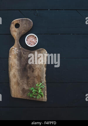 Cucina-ware set. Vecchio rustico tagliere realizzato in legno di noce, sale e origano herb su uno sfondo nero, vista dall'alto, copia spa Foto Stock