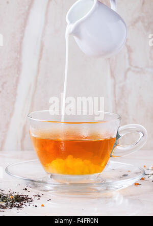 Versare il latte dalla caraffa del bianco al vetro tazza di tè caldo sul piattino a secco con verde e nero le foglie di tè su marmo bianco backgtound. Foto Stock