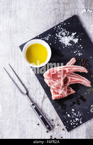 Materie costolette di agnello con sale e pepe, asciugare le erbe aromatiche e la coppa dell' olio d' oliva sul nero ardesia bordo bianco su un tavolo di legno. Forcella di carne vicino a. Foto Stock