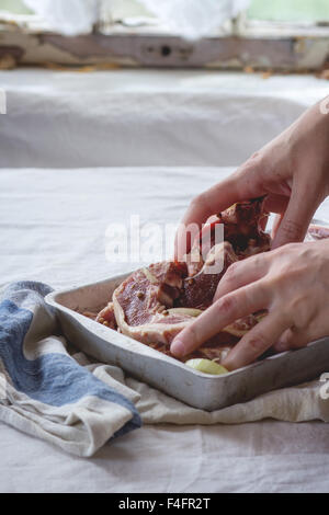 Donna di agitazione manuale crudo marinato costolette di agnello con la cipolla e il pepe in alluminio vecchia teglia. Su tavolo da cucina con il bianco Foto Stock