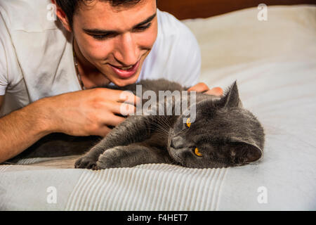 Bel giovane uomo Animal-Lover su un letto, avvolgente e coccole il suo grigio gatto domestico Pet. Foto Stock