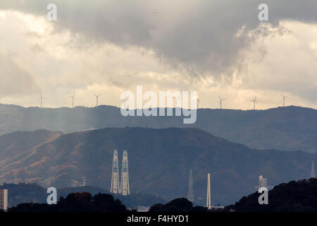 Wakayama, Giappone, stagliano le turbine eoliche sulla cima delle montagne in lontananza con vari camini industriali in primo piano. Tempesta di pioggia, ma alcuni la luce del sole. Foto Stock