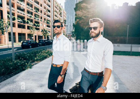 Ginocchio la figura dei due giovani barbuto bionda e capelli neri imprenditore moderno, passeggiate nella città di retroilluminazione, entrambi affacciati a destra - lavoro, concetto di successo Foto Stock