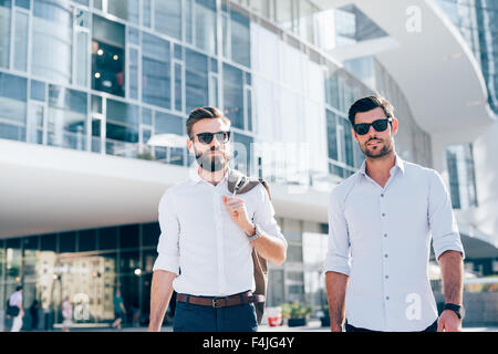 A metà lunghezza di due giovani barbuto bionda e capelli neri imprenditore moderno, passeggiate nella città di retroilluminazione, uno che guarda in telecamera, l'altro che si affaccia a destra - lavoro, concetto di successo Foto Stock