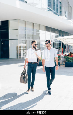 Due giovani barbuto bionda e capelli neri imprenditore moderno, passeggiate nella città di retroilluminazione, uno affacciato sulla destra, l'altro che si affaccia a sinistra - lavoro, concetto di successo Foto Stock