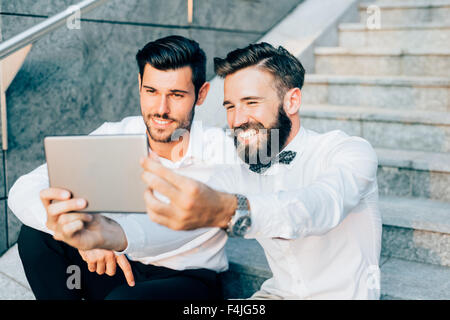 Due giovani barbuto bionda e capelli neri imprenditore moderno, seduti su una scalinata, utilizzando una compressa, prendendo un selfie - tecnologia, vanità, sociale un concetto di rete Foto Stock