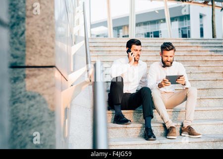 Due giovani barbuto bionda e capelli neri imprenditore moderno, seduti su una scalinata, che parla di smartphone e tablet utilizzando - tecnologia, business, il concetto di lavoro Foto Stock
