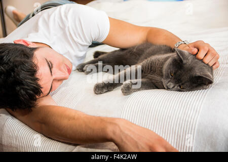 Bel giovane uomo Animal-Lover su un letto, avvolgente e coccole il suo grigio gatto domestico Pet. Foto Stock