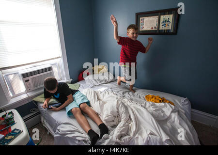 Un giovane ragazzo guarda al suo tablet mentre il suo fratello più giovane fastidiosamente danze sul suo letto Foto Stock