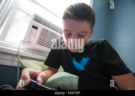 Un giovane ragazzo guarda al suo tablet da sola nella sua camera da letto in una giornata di sole Foto Stock