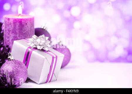 Viola e argento baubles di Natale, un dono e una candela di fronte defocalizzata viola e luci bianche. Foto Stock