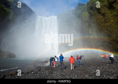 I turisti al rainbow al di sotto di 60m-alta cascata Skogafoss, Skogar, Sudhurland, Islanda. Foto Stock