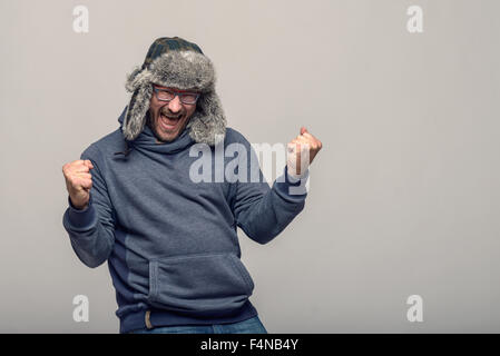 Felice l'uomo con gli occhiali e un cappello invernale il tifo e celebra il suo sollevamento rinserrata pugni in aria con un esultante expre Foto Stock